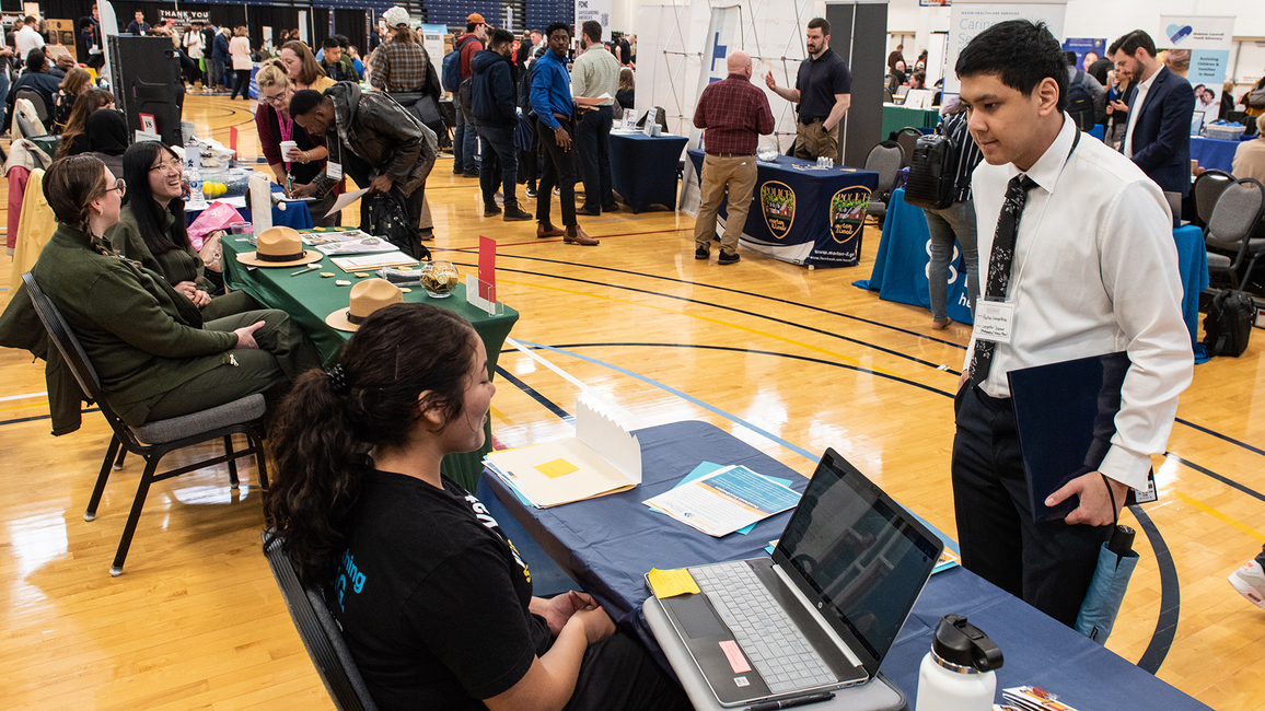 A man stands in front of a table, speaking to a woman who is seated at the 2023 Career Connections Expo.